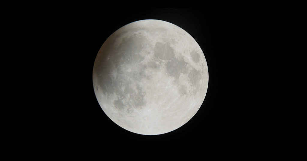 Photo montrant la pleine lune avec une petite zone éclipsée à gauche du globe, dont on devine qu'elle est légèrement ocre. Le fond de ciel est noir.